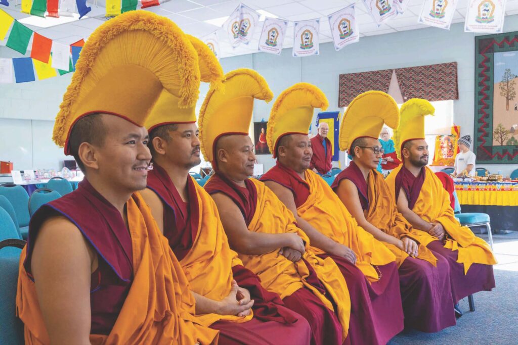 Tibetan Buddhist monks sit in a row. They are wearing red robes and yellow hats