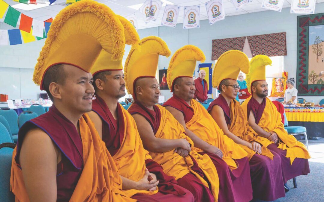 Tibetan Buddhist monks sit in a row. They are wearing red robes and yellow hats