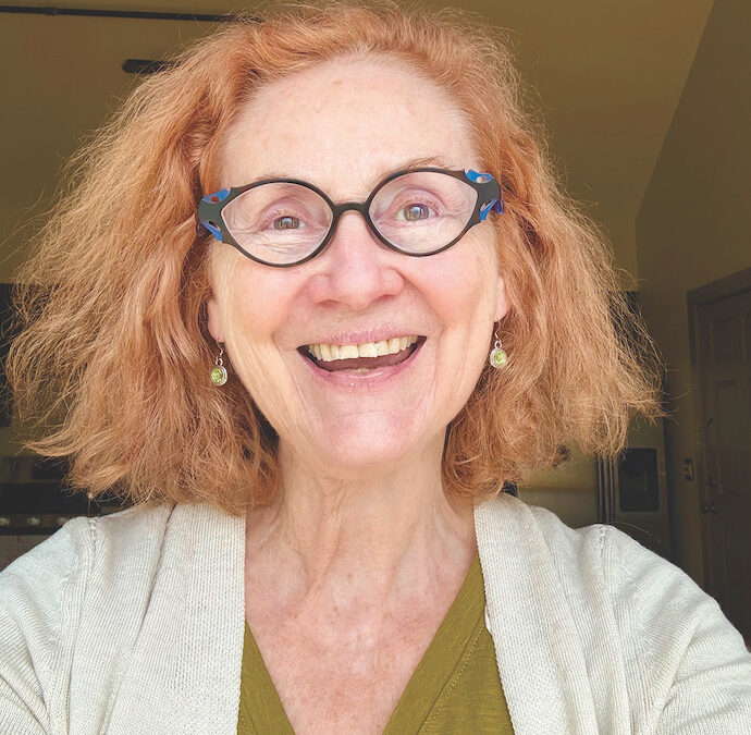 A woman with blue glasses and red hair smiles at the camera.