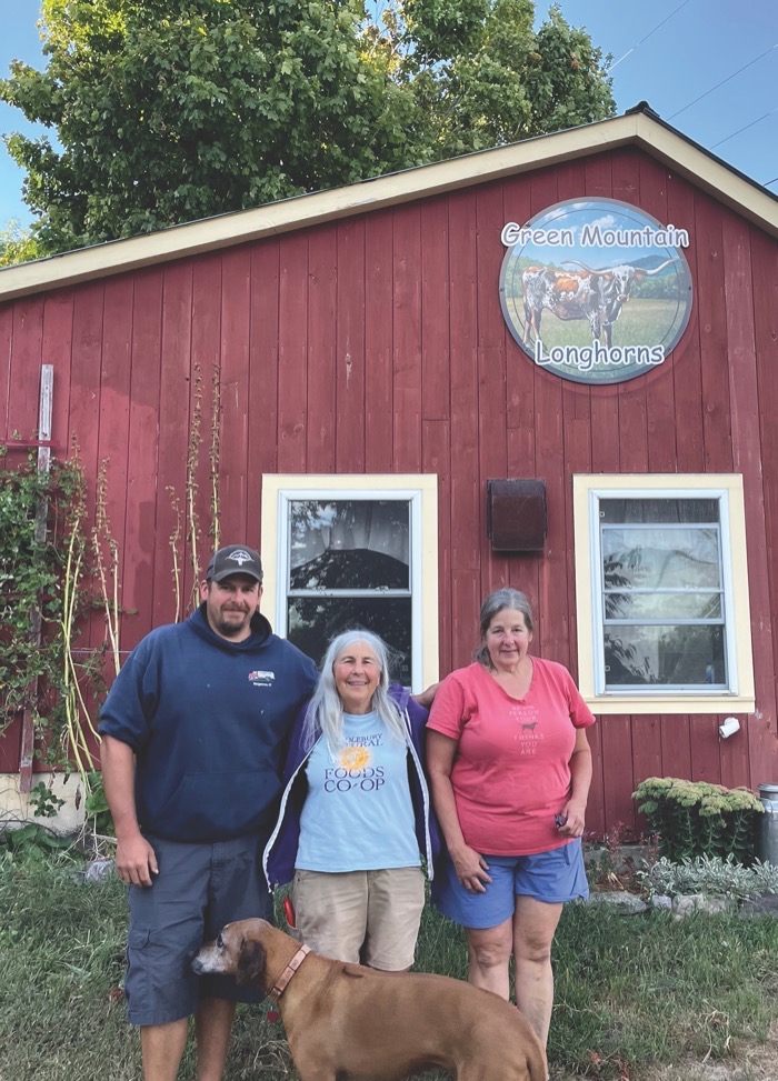 2 women, 1 man, and a dog stand in front of a red barn and a sign reading 'Green Mountain Longhorns.'