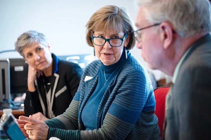 A middle-aged white woman woman in a turquoise and grey striped sweater and turquoise glasses converses with a man.