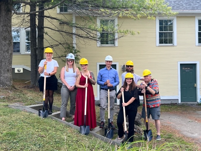 People in hardhats hold shovels and stand in front of a yellow building.